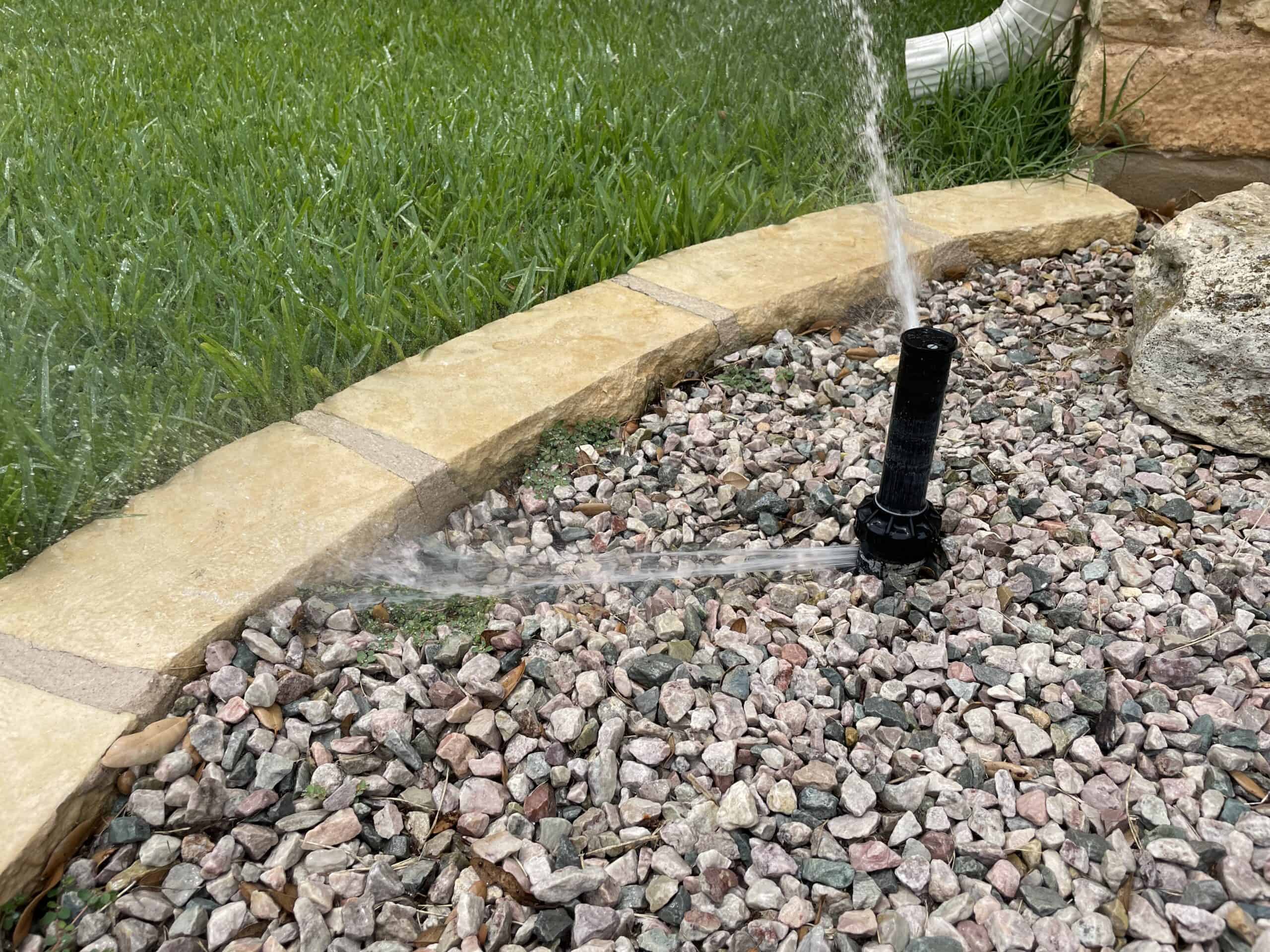 Lawn sprinkler spraying water in multiple directions, installed in a stone-bordered mulch bed next to a green, well-maintained lawn.