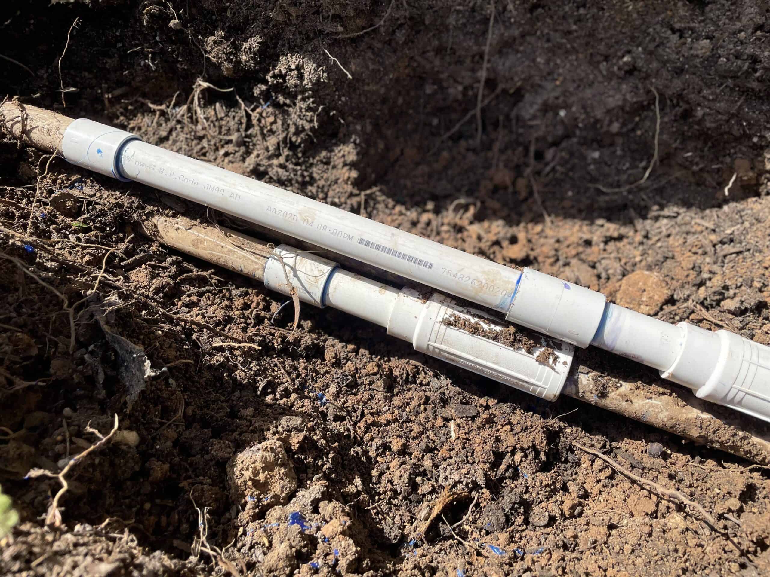Two white PVC irrigation pipes exposed in a trench, showing pipe couplings and connections prepared for underground water line repair.