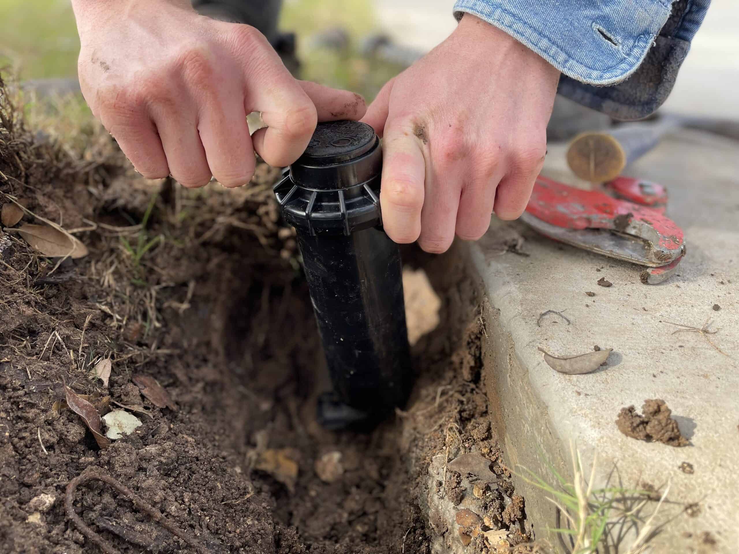 Hands gripping a black sprinkler head, performing maintenance near a concrete edge with a red pipe cutter and a hammer in the background.