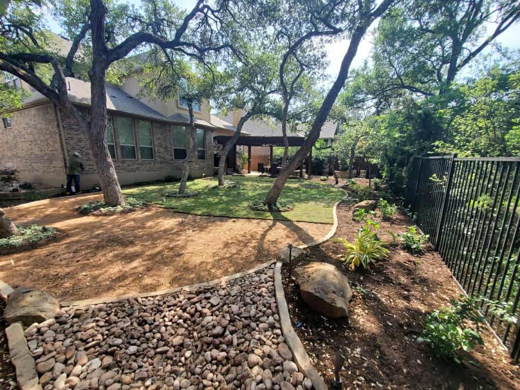 Shaded backyard with mature trees, curved pathways, stone borders, and landscaped garden beds surrounding a grassy entertaining space.