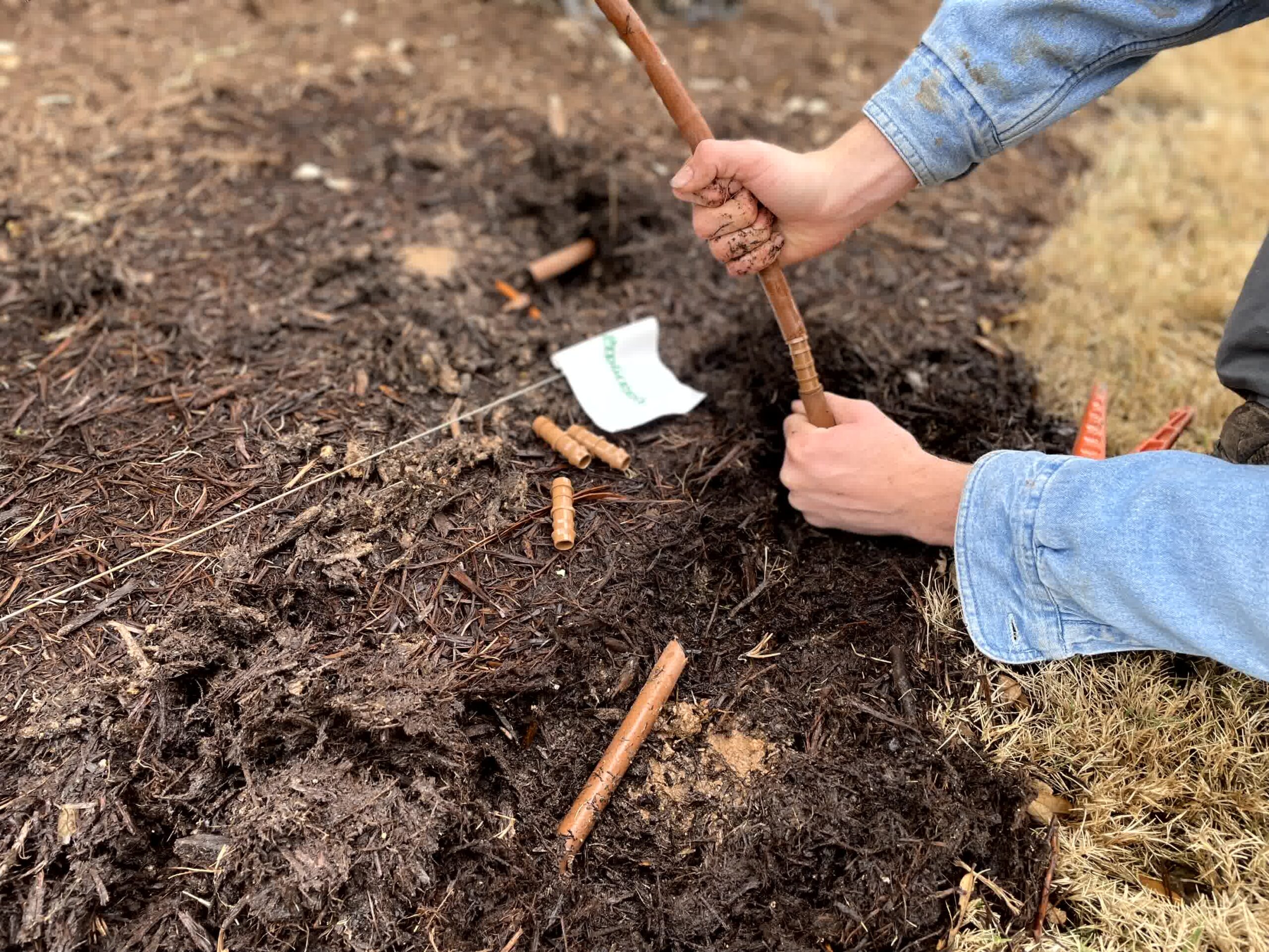 Technician connects brown drip irrigation tubing using barbed fitting in mulch bed, with ProGreen flag marking repair area.