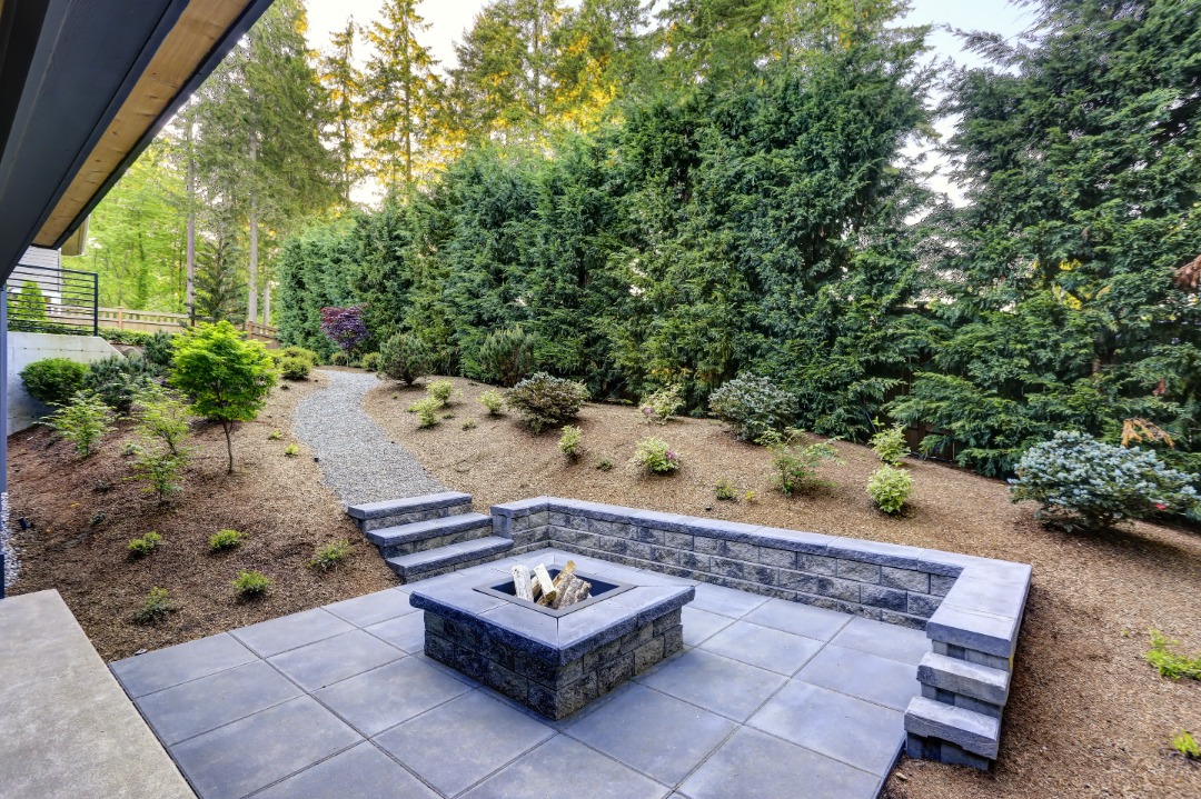 An outdoor living space with a firepit and tall green trees in the background.

