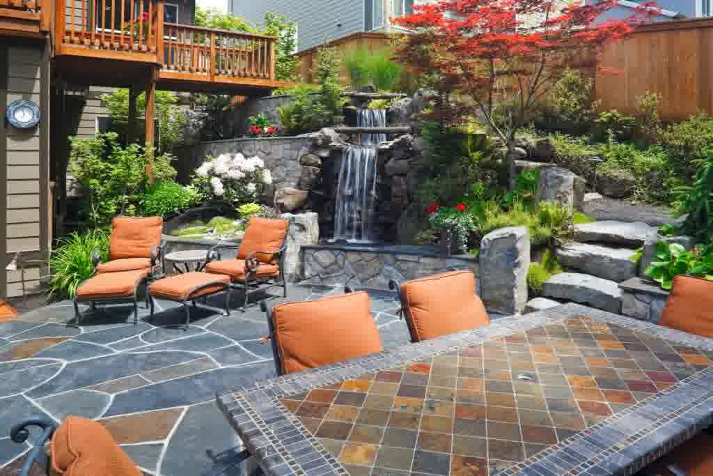 A luxurious outdoor living room with chairs, a waterfall, a table, a stone staircase, and green foliage.
