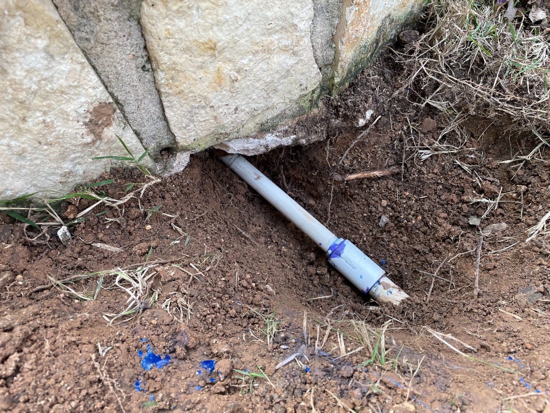 irrigation pipe runs beneath stone retaining wall with visible blue sealant on joint, showing underground water line connection.
