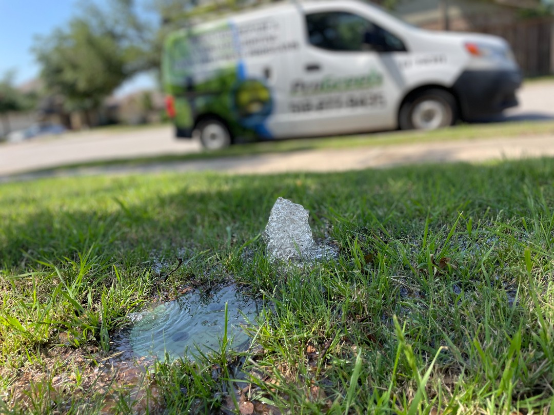 Leaking irrigation head sprays water into a small puddle on a lawn with a blurred landscaping van in the background.