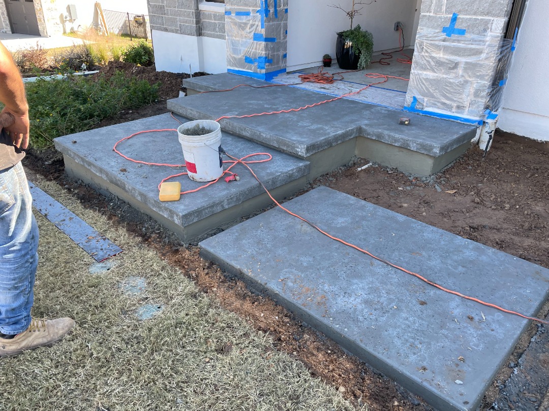 Freshly poured concrete steps lead to a home's front porch, surrounded by tools and materials for ongoing landscape work.