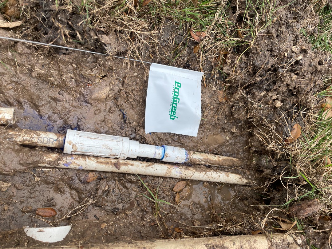 Exposed PVC irrigation pipes in muddy trench show recent repair work with fittings and ProGreen flag marking irrigation zone.