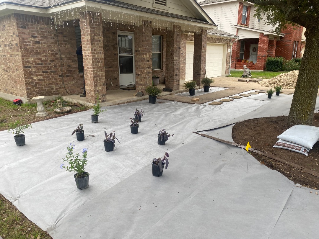 Front yard landscape installation with weed barrier fabric and potted plants arranged for a clean and organized garden layout.