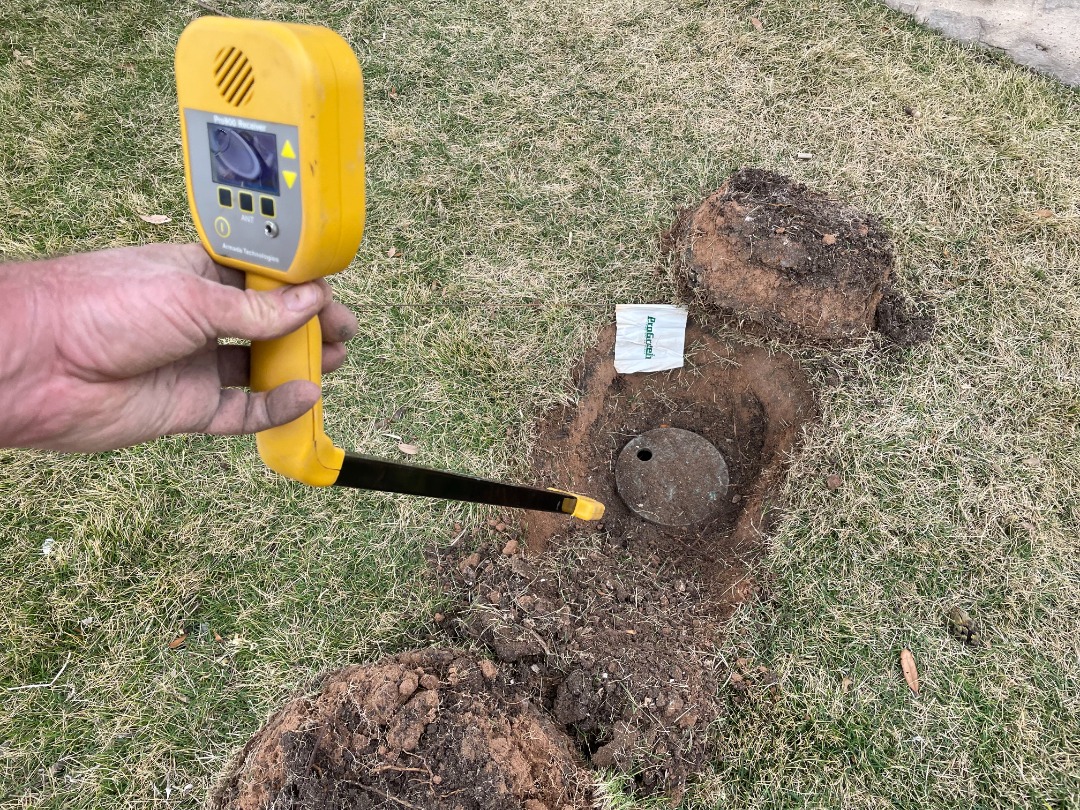 Technician uses a yellow pipe locator to detect a buried irrigation valve in a dug-out lawn section with exposed soil and a small ProGreen marker flag.