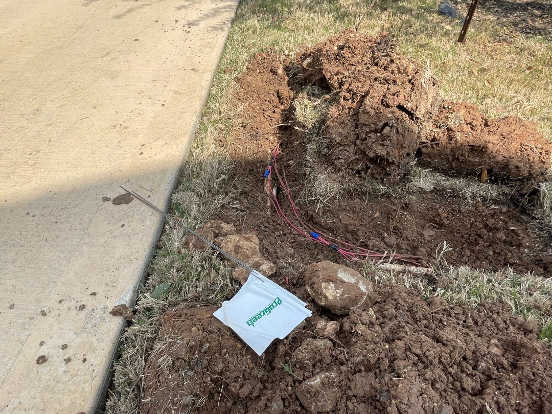 Exposed irrigation control wires beside a concrete driveway during trenching, marked with a ProGreen flag and surrounded by dug-up soil.