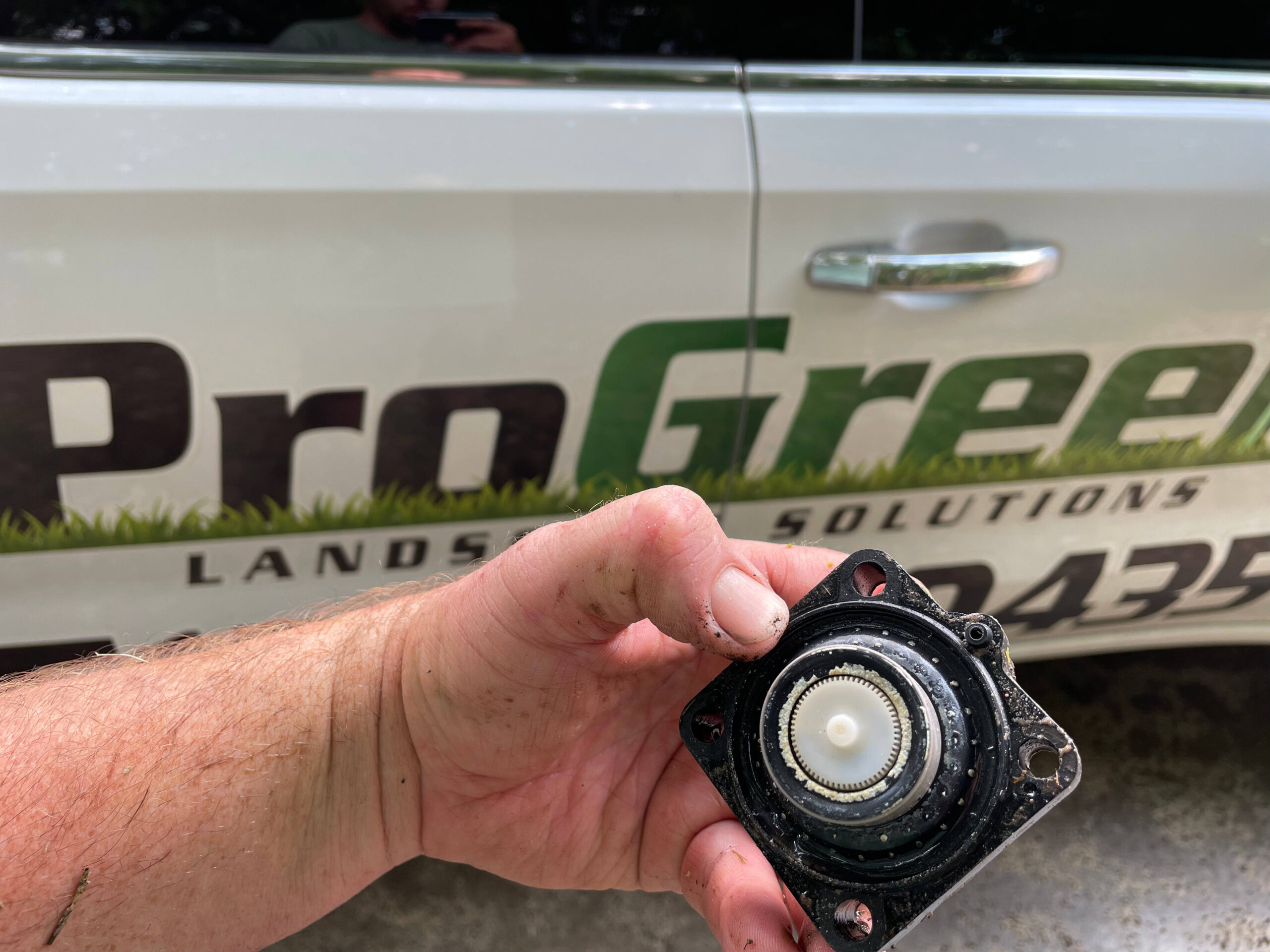 Close-up of technician holding irrigation valve diaphragm assembly with exposed gears in front of a ProGreen Landscapes service vehicle.
