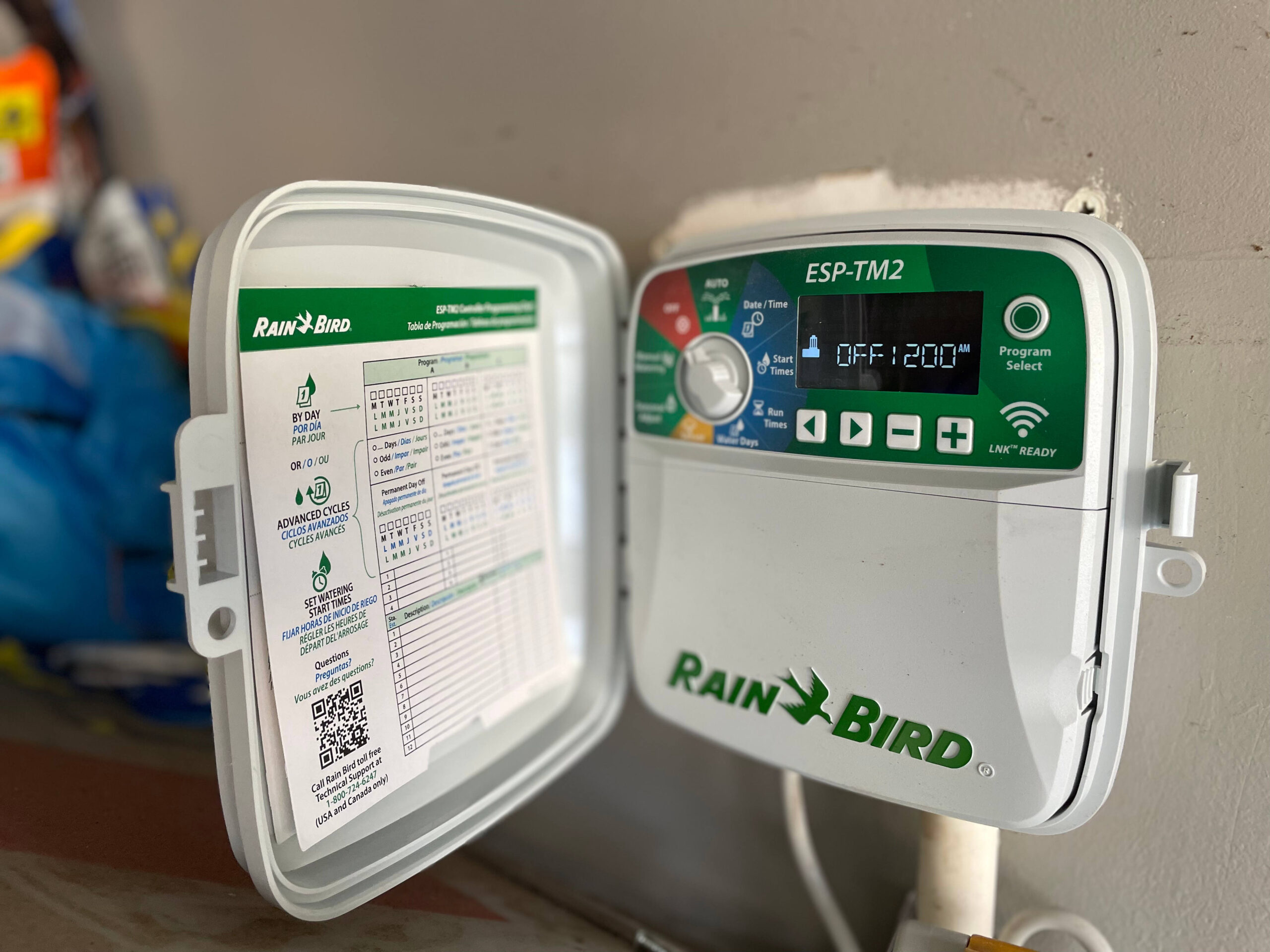 Rain Bird ESP-TM2 irrigation timer with open programming guide, set to off, mounted on interior wall with wires and tubing visible.