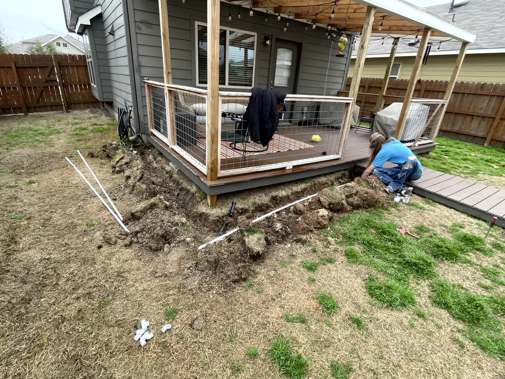 Backyard trenching project in progress with exposed PVC piping and person installing irrigation line next to wooden deck.