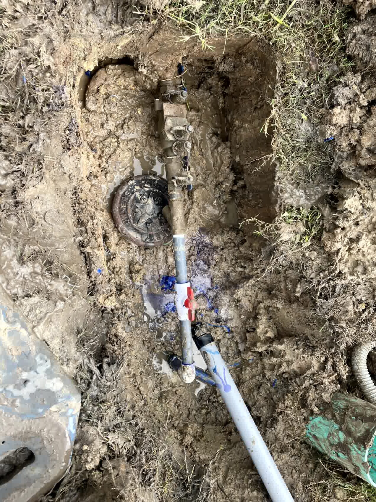 Broken irrigation line and valve exposed in muddy soil with pooling water, showing system leak before repair.