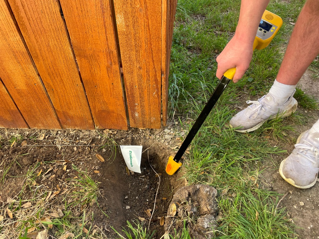 Technician uses a pipe locator to identify an underground irrigation line near a wooden fence, marking the area for repair.